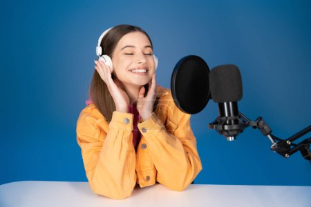 Pleased teenager in headphones sitting near studio microphone isolated on blue 