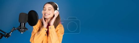 Joyful teenager in headphones standing near studio microphone isolated on blue, banner 