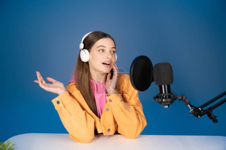 Teen girl in headphones talking during podcast near studio microphone isolated on blue 