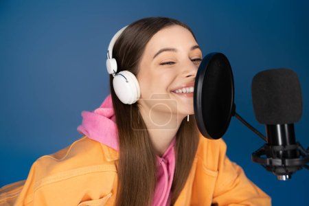 Cheerful teenager in headphones talking near studio microphone during podcast isolated on blue 