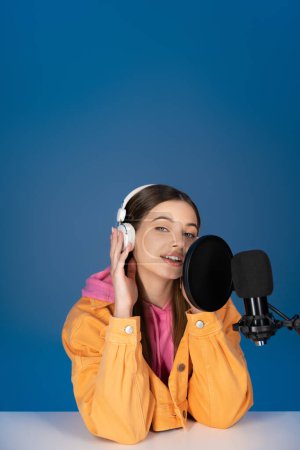 Brunette teenager touching headphones and talking near studio microphone isolated on blue 