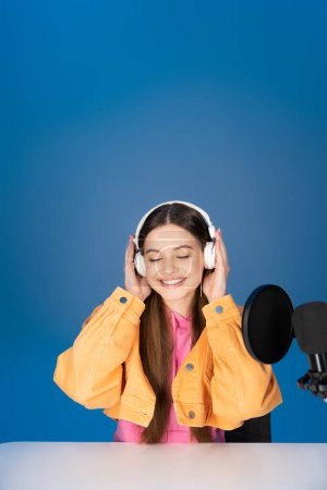 Cheerful brunette teenager in headphones sitting near microphone and table isolated on blue 