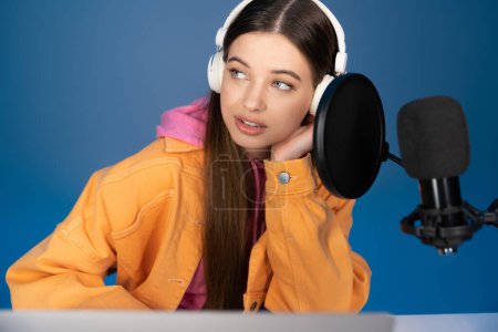 Teenager in headphones looking away near studio microphone isolated on blue 