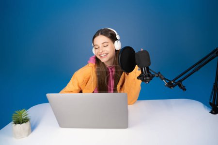 Smiling teen girl in headphones using laptop near microphone on table isolated on blue 
