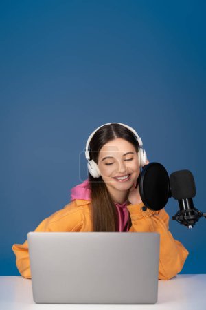 Smiling teenage girl in headphones looking at laptop near studio microphone isolated on blue 