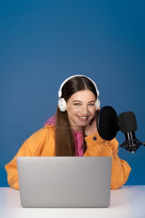 Téléchargez les photos : Brunette teen girl in headphones looking at camera near laptop and microphone isolated on blue - en image libre de droit