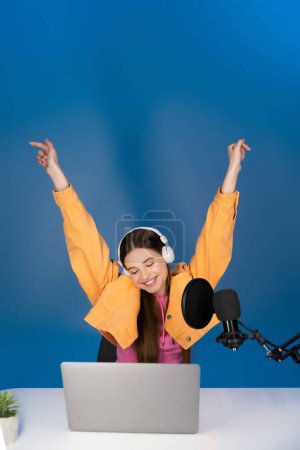 Pleased teenager in headphones sitting with raised hands near microphone and laptop on blue background 