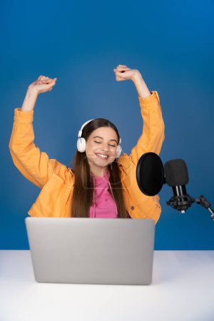 Excited teenager in headphones using laptop and studio microphone and sitting with raised hands isolated on blue 