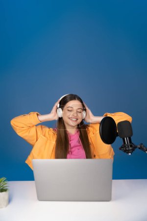 Smiling teen girl in headphones using laptop near microphone isolated on blue 