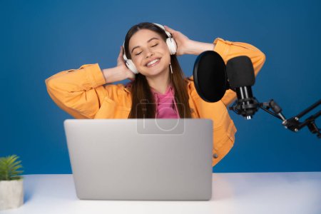 Happy teenager touching headphones near laptop and studio microphone isolated on blue 