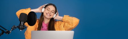 Teenager with closed eyes touching headphones near laptop and studio microphone isolated on blue, banner 