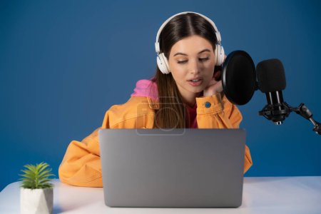 Brunette teenager in headphones talking near studio microphone and laptop on table isolated on blue 