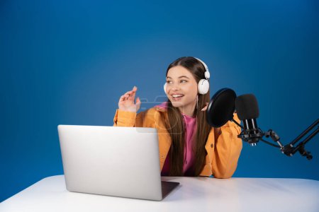 Teen girl in headphones recording podcast near studio microphone and laptop isolated on blue 
