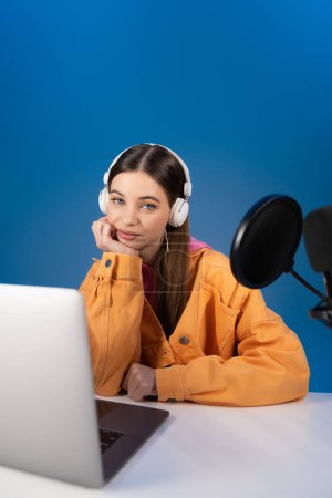 Téléchargez les photos : Teen brunette girl in headphones looking at camera near blurred laptop and studio microphone isolated on blue - en image libre de droit