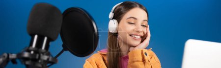 Cheerful teenager in headphones doing podcast near laptop and studio microphone isolated on blue, banner 