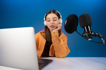 Téléchargez les photos : Teenager in headphones looking at laptop while recording podcast isolated on blue - en image libre de droit