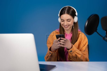 Smiling teenager in headphones using smartphone near laptop and microphone isolated on blue 