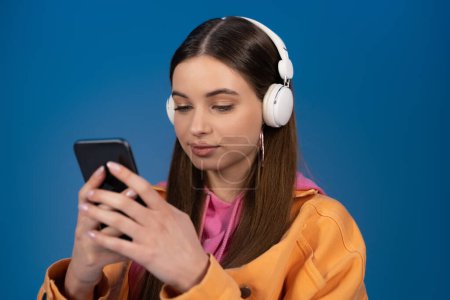Teen girl in wireless headphones chatting on smartphone isolated on blue 