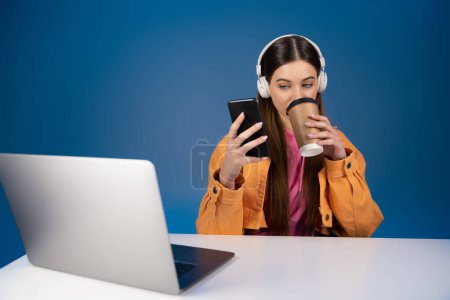 Teenager in headphones using smartphone and drinking takeaway coffee near laptop isolated on blue 