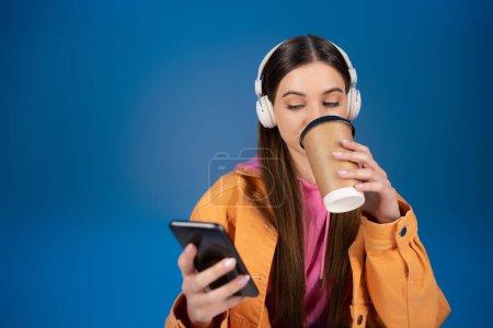 Photo for Teenage girl in headphones chatting on smartphone and drinking coffee to go isolated on blue - Royalty Free Image
