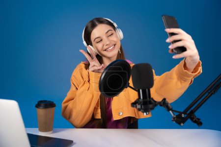 Teenager in headphones taking selfie on smartphone near studio microphone and laptop isolated on blue 