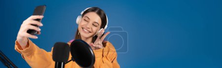 Téléchargez les photos : Teenager in headphones taking selfie on smartphone near studio microphone isolated on blue, banner - en image libre de droit