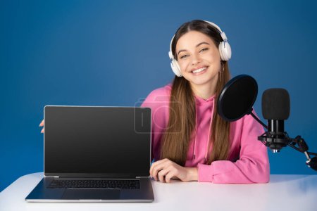 Smiling teenager in headphones looking at camera near laptop with blank screen and studio microphone isolated on blue 