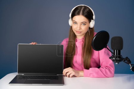 Foto de Teen podcaster in headphones sitting near laptop and microphone isolated on blue - Imagen libre de derechos