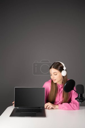 Teen podcaster looking at laptop with blank screen near microphone isolated on grey 
