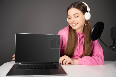 Téléchargez les photos : Smiling teen podcaster looking at laptop with blank screen near microphone isolated on grey - en image libre de droit