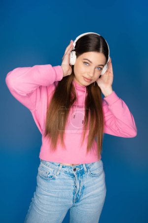 Teenager in hoodie listening music in headphones isolated on blue 