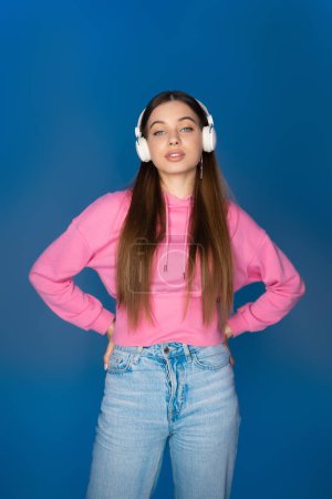 Foto de Teen girl in jeans and hoodie listening music in headphones isolated on blue - Imagen libre de derechos