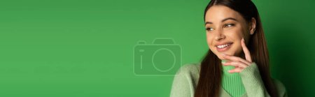 Foto de Positive teenage girl in jumper looking away on green background, banner - Imagen libre de derechos