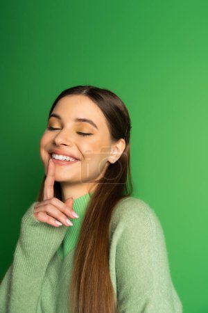 Smiling teenager in jumper touching lip on green background 