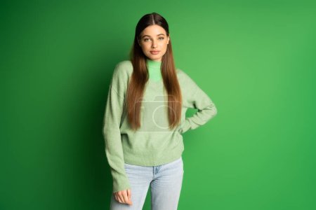 Brunette teenager in jeans and jumper holding hand on hip on green background 