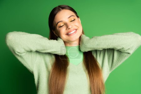 Foto de Joyful teenage girl in jumper touching neck on green background - Imagen libre de derechos