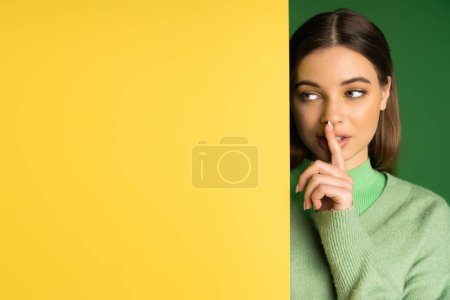 Brunette teen girl showing shh sign near yellow and green background 