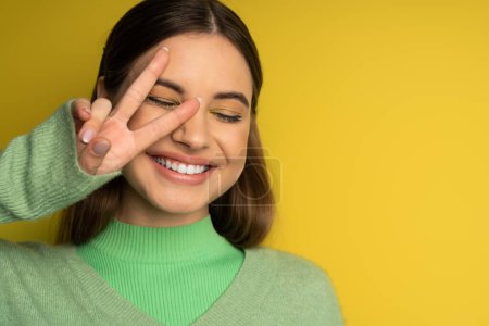 Positive teen girl in jumper showing peace sign on yellow background 
