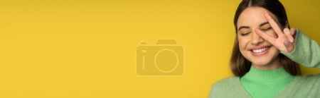 Foto de Cheerful brunette teen girl showing peace gesture on yellow background, banner - Imagen libre de derechos