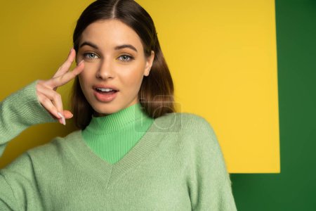 Teen girl in jumper showing victory sign on yellow and green background 