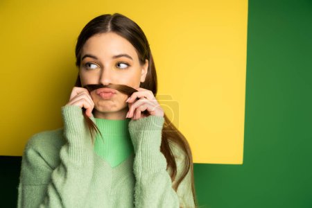 Brunette teen girl holding hair near lips on yellow and green background 