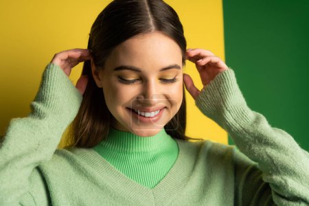 Foto de Teen girl in jumper touching hair and smiling on green and yellow background - Imagen libre de derechos