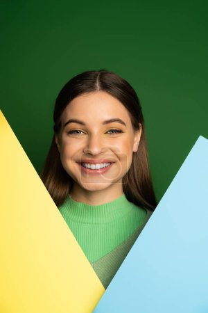 Smiling teenager with makeup looking at camera near colorful paper isolated on green 