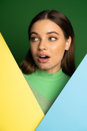 Foto de Shocked teen girl with visage looking away near colorful paper isolated on green - Imagen libre de derechos