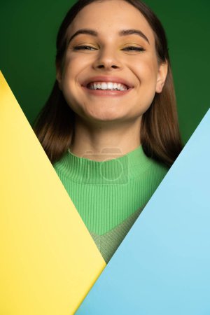 Positive teen girl with visage looking at camera near colorful paper isolated on green 
