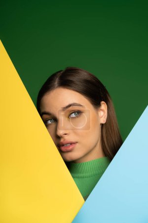 Brunette teenager in turtleneck posing near colorful paper isolated on green 