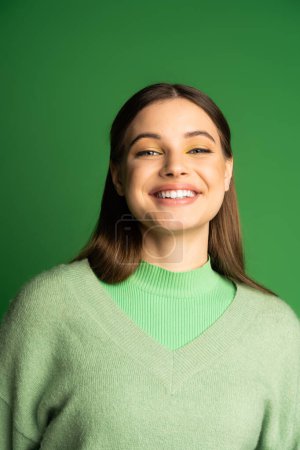 Photo for Portrait of happy teen girl in jumper smiling at camera isolated on green - Royalty Free Image