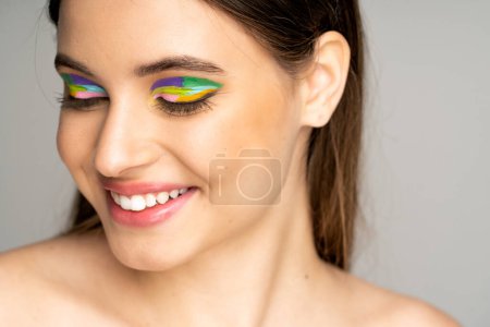 happy teen model with colorful makeup smiling isolated on grey 
