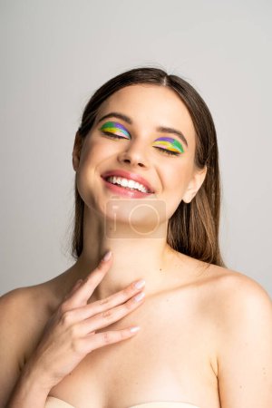 Positive teen model with colorful visage touching neck isolated on grey 