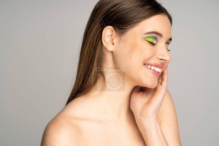 Smiling teen model with colorful visage and naked shoulders touching face isolated on grey 